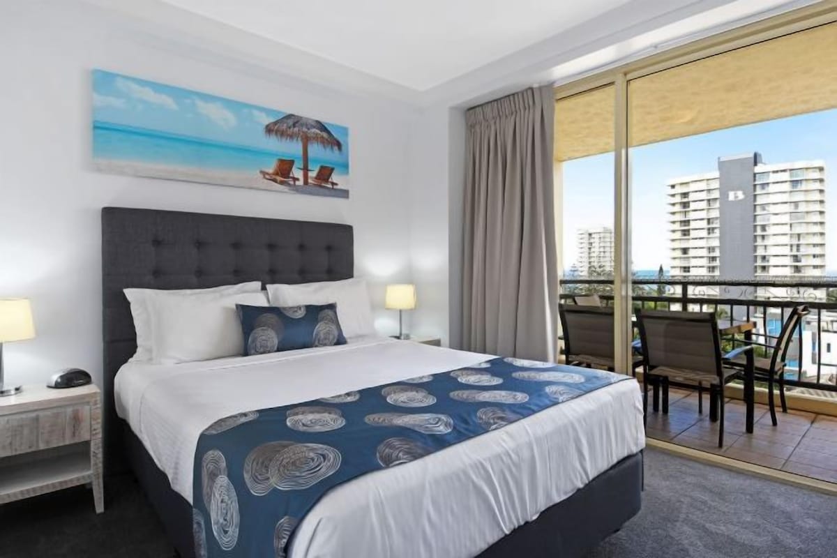 A hotel room with a large bed, a painting of a beach scene, a balcony with chairs, and a city view.