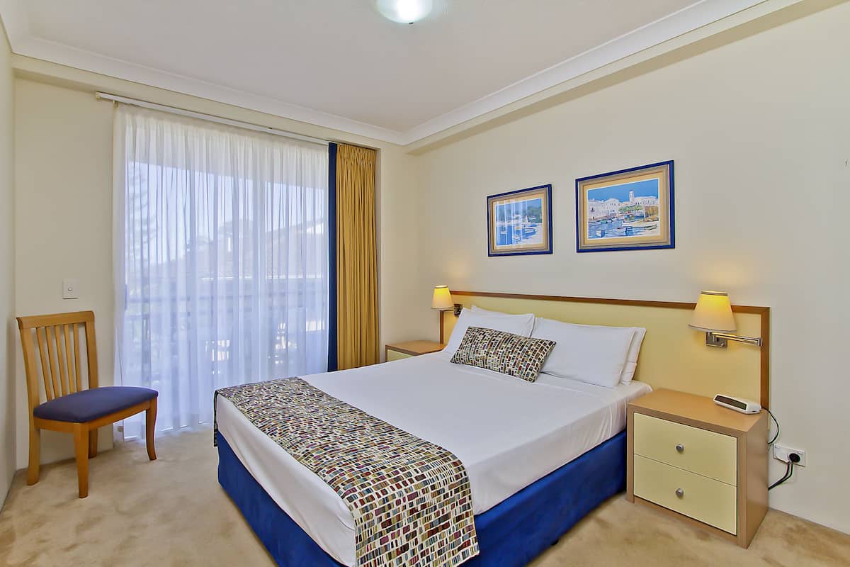 Una habitación de hotel con cama, una silla, una mesita de noche y dos cuadros en la pared.