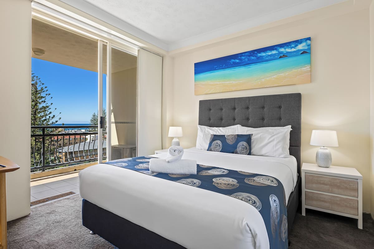 Habitación de hotel con una cama grande, dos mesitas de noche con lámparas, un cuadro de una playa y un balcón con vistas a la ciudad.