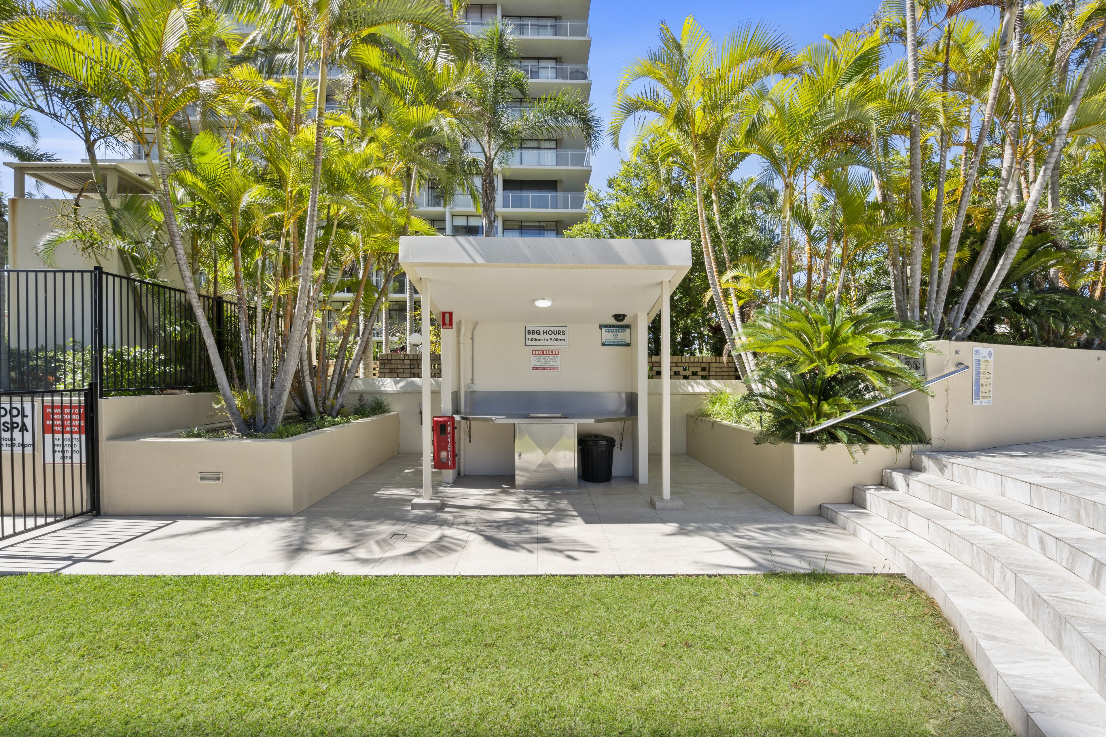 A covered outdoor area with a BBQ grill, surrounded by palm trees and a building in the background.