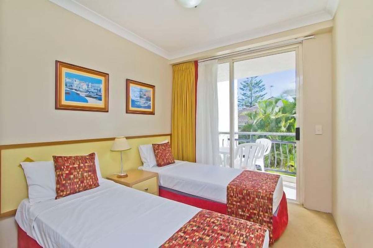A hotel room with two beds, a balcony with a view of greenery, and two framed pictures on the wall.