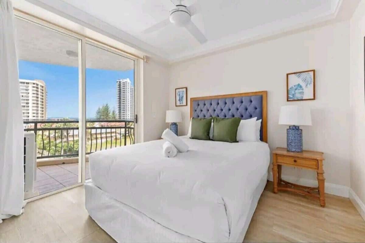 Un dormitorio con una cama grande, una mesita de noche, un ventilador de techo y vistas a edificios a través de una puerta corrediza de vidrio.