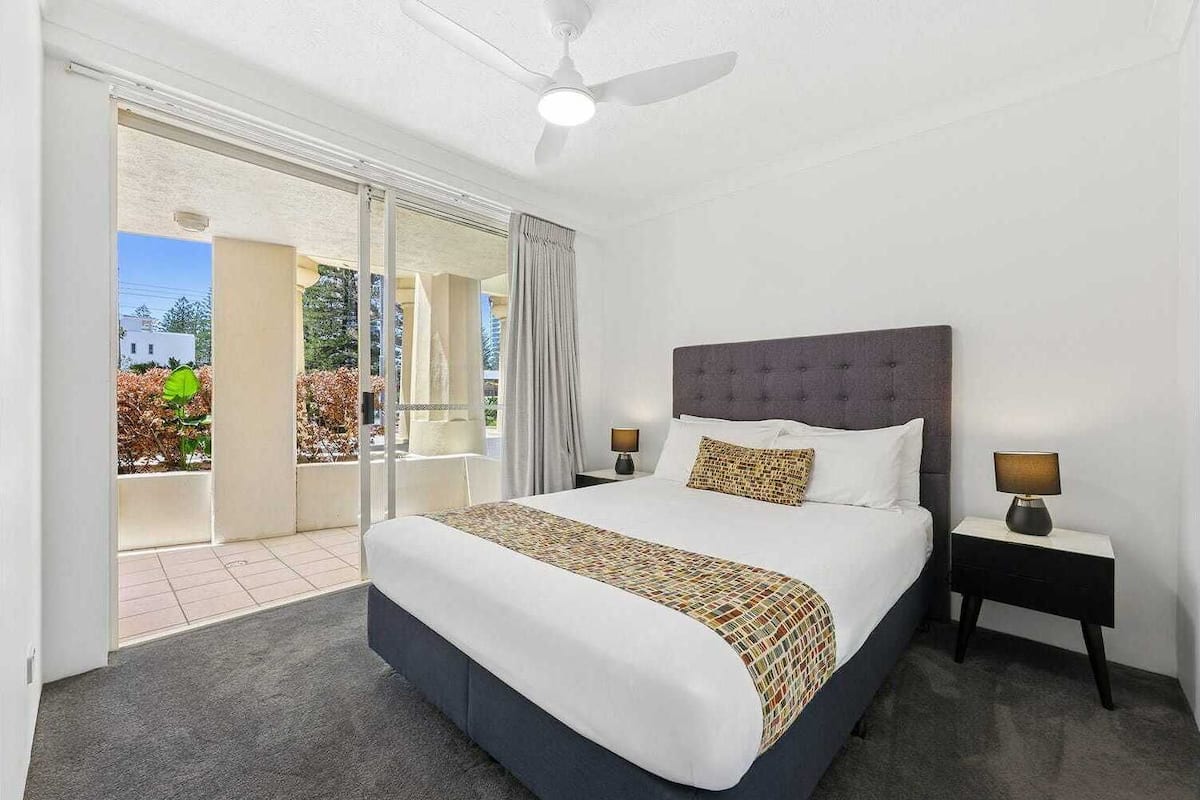 Habitación de hotel con una cama grande, un ventilador de techo, una mesita de noche con lámpara y un ventanal corredero que da a un balcón.