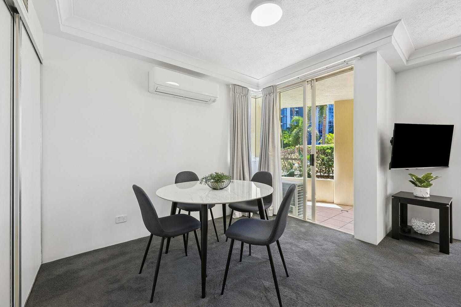 A room with a round table, four chairs, a small potted plant, a TV on a stand, and a sliding glass door leading to an outdoor area.