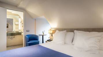 A hotel room with a bed, a blue chair, a small table, and a bathroom visible through an open door.