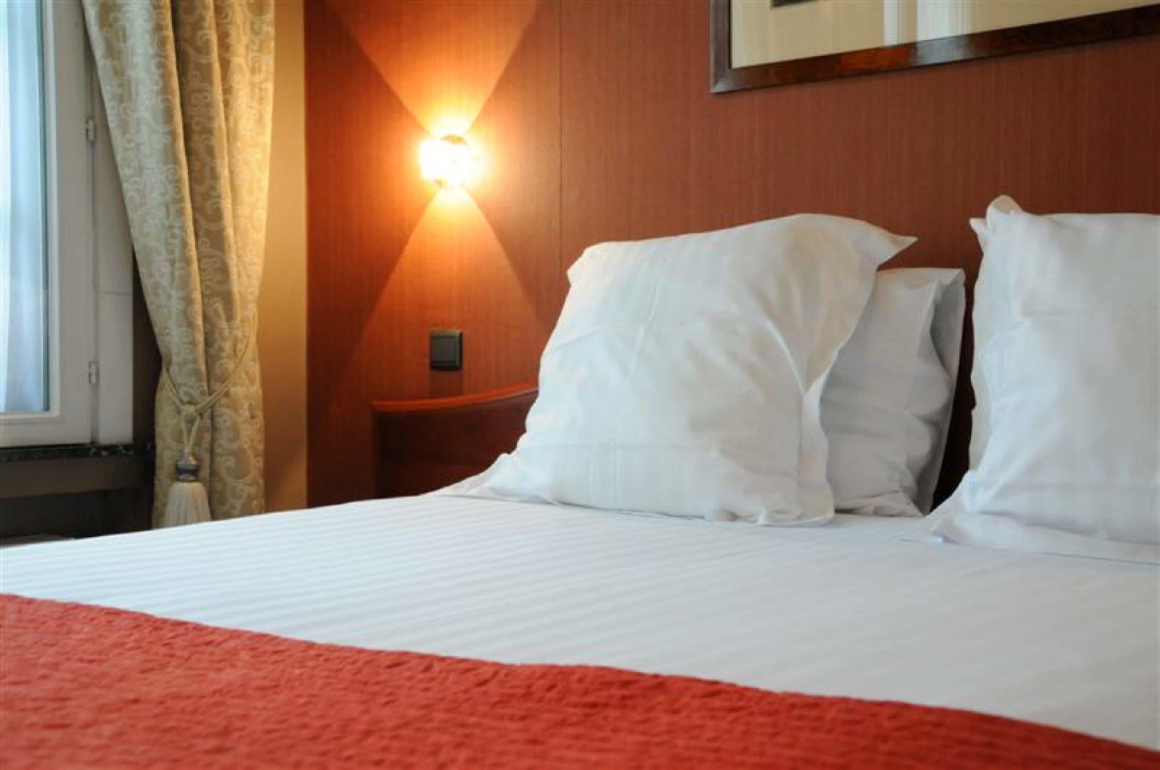 Habitación de hotel con una cama, almohadas blancas, una colcha roja, una lámpara de pared y un ventanal con cortinas.