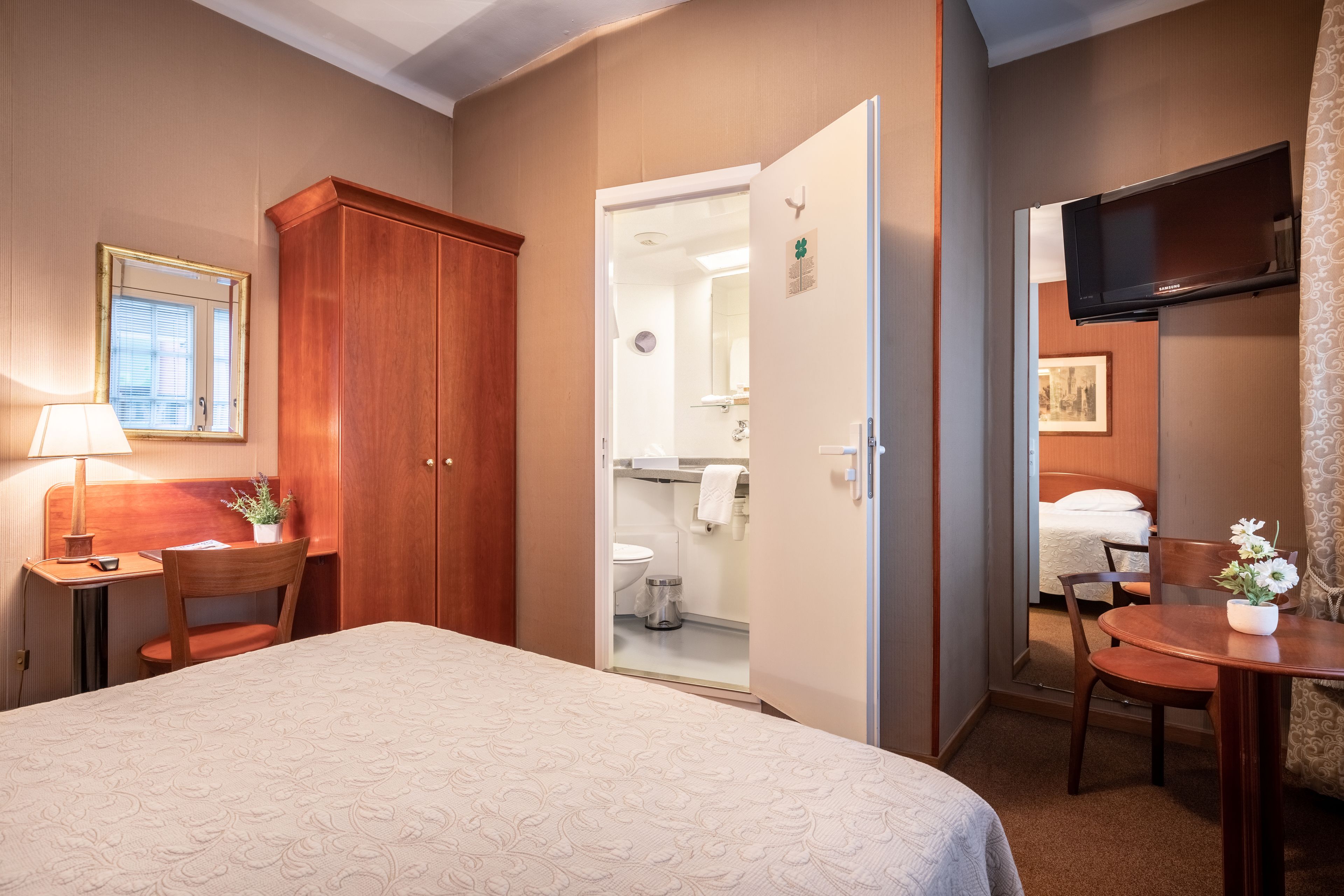 A hotel room with a bed, a desk with a lamp, a wardrobe, a bathroom visible through an open door, and a TV mounted on the wall.