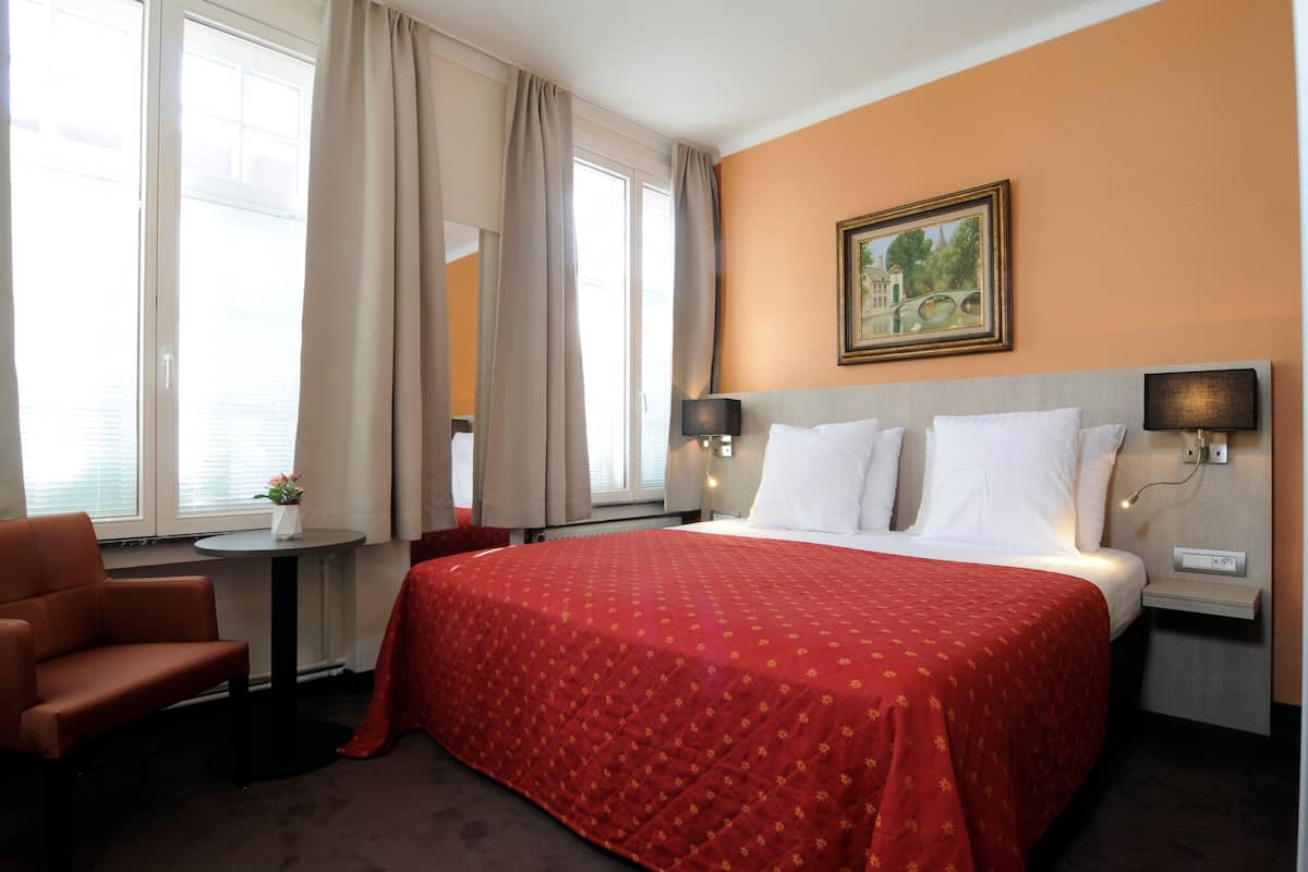 Hotellihuone, jossa on suuri sänky, tuoli, pieni pöytä ja taulu seinällä.