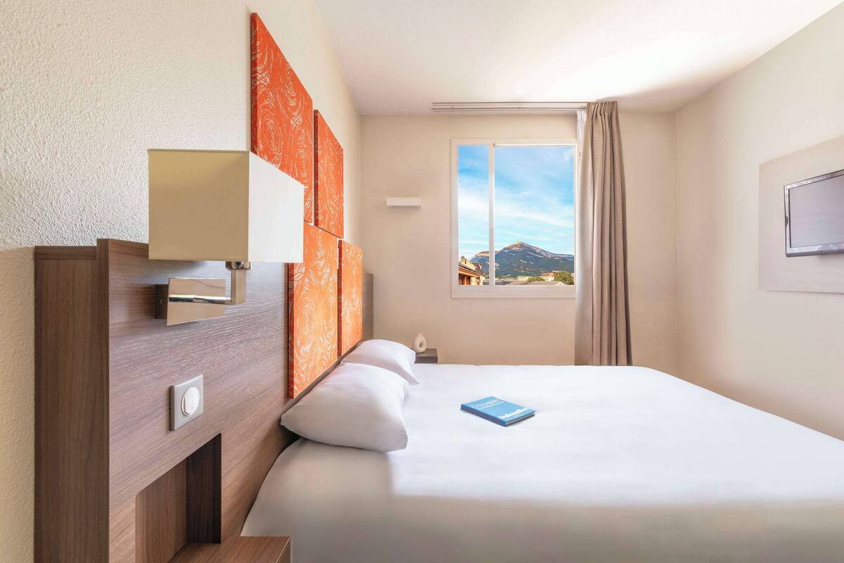 Habitación de hotel con cama, mesita de noche, lámpara, ventana con vista a montañas y televisor montado en la pared.