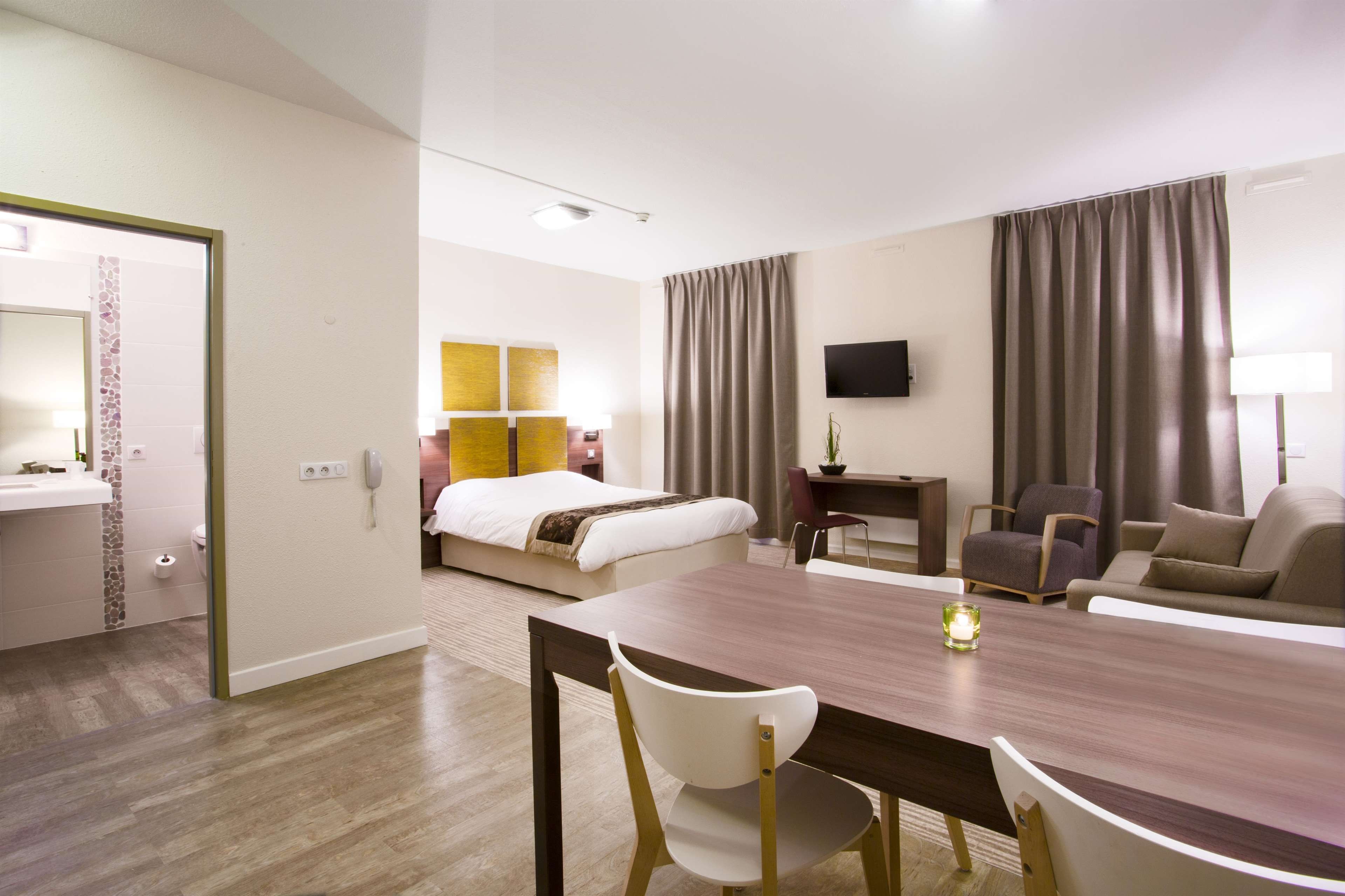 A hotel room with a bed, a desk, a TV, a dining table with chairs, and a bathroom with a mirror and sink.