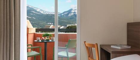 A hotel room with a bed, a wooden chair, a small table with a book, and a balcony with chairs and a table.