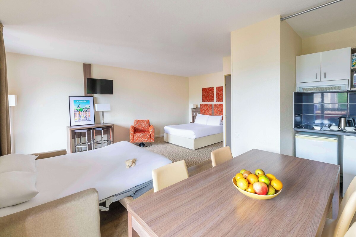 Habitación de hotel con dos camas, una mesa de comedor con fruta, un televisor y una kitchenette.