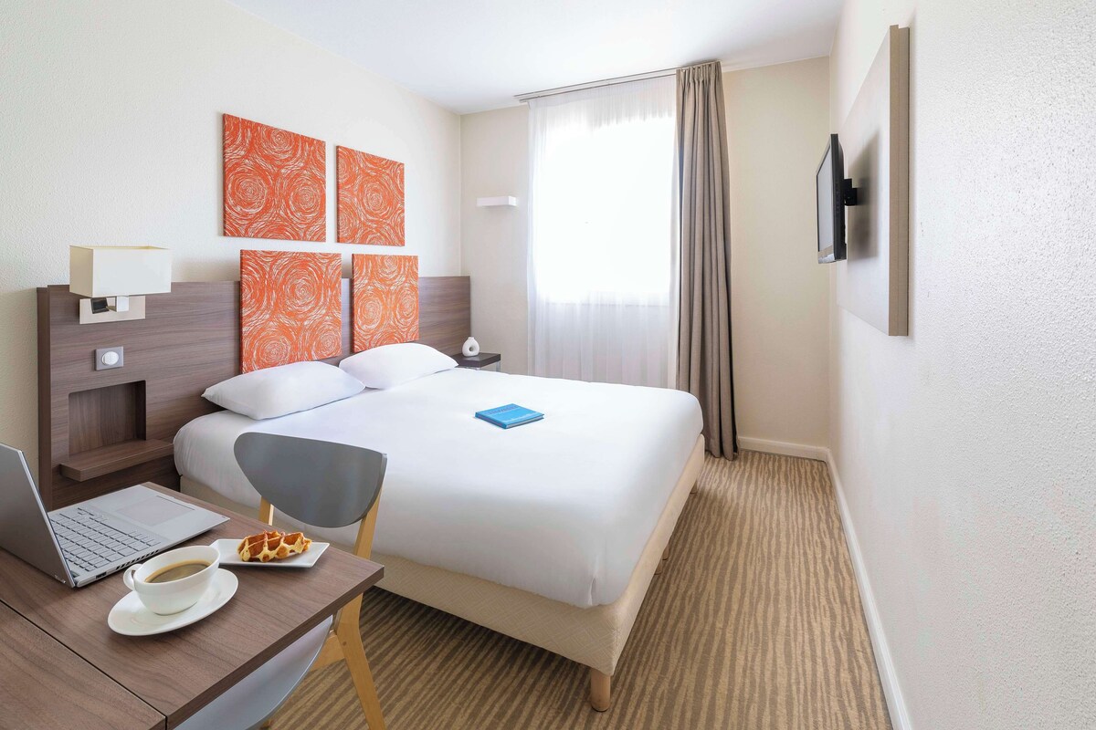 Una habitación de hotel con una cama, un escritorio con una computadora portátil y una taza de café, una silla y un televisor montado en la pared.