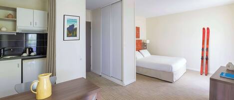 Habitación de hotel con una cama, un escritorio con silla, una kitchenette y un par de esquíes apoyados en la pared.