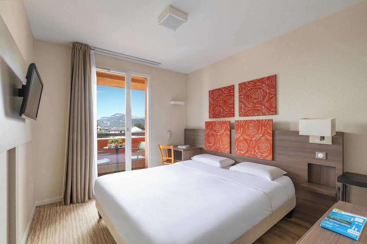 Habitación de hotel con cama, escritorio, silla, balcón con vista a montañas y obras de arte en la pared.