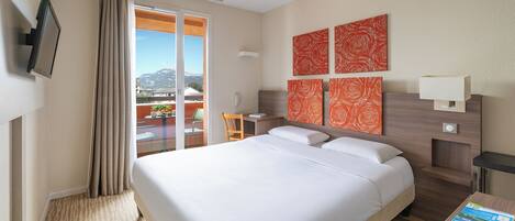 A hotel room with a bed, a desk, a chair, a balcony with a view of mountains, and artwork on the wall.