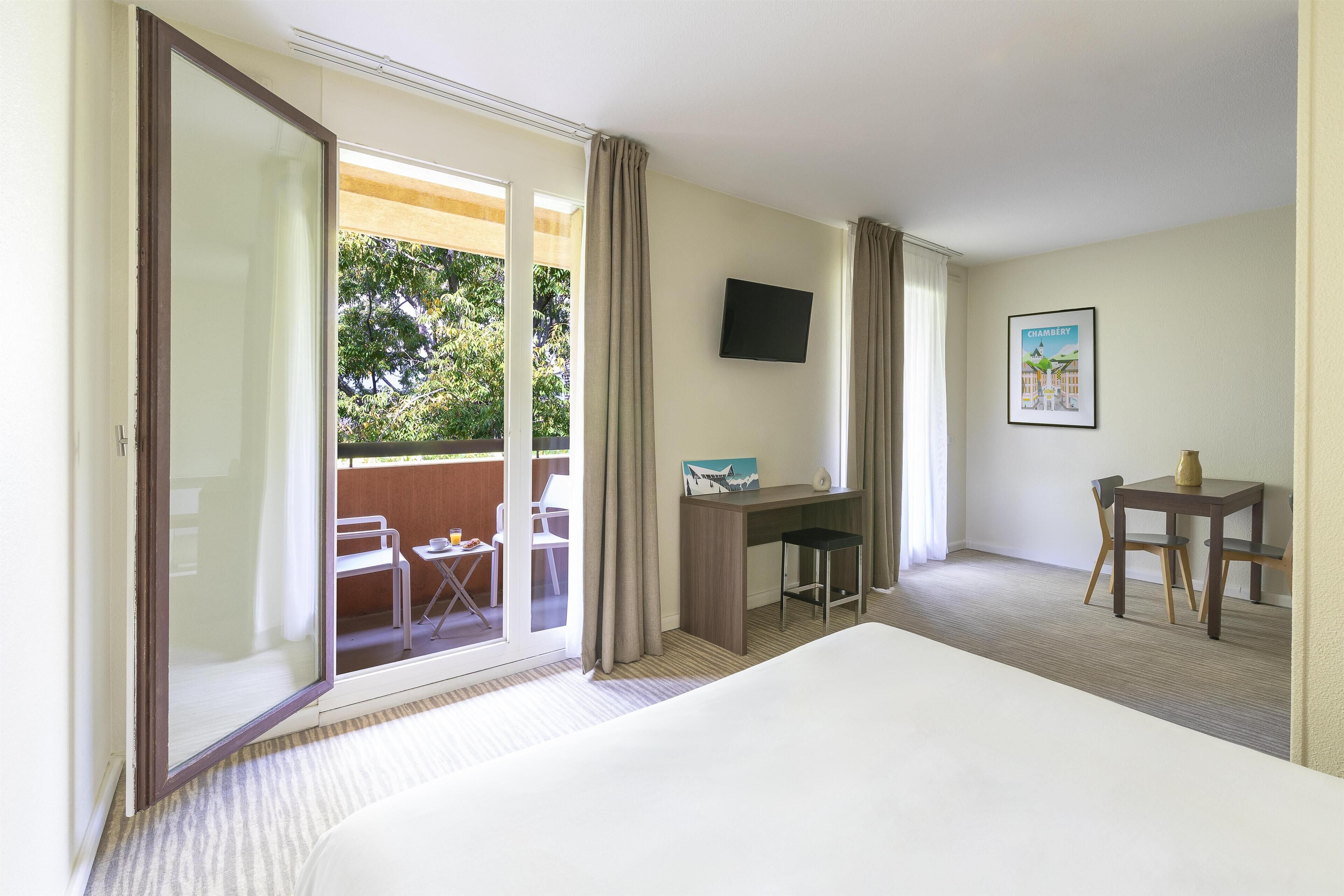 A hotel room with a bed, a desk, a chair, a TV, and a balcony with outdoor furniture.