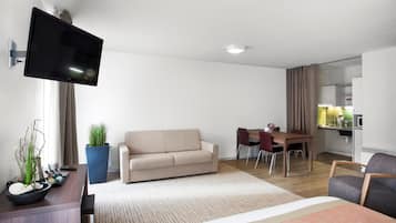 A modern hotel room with a flat-screen TV mounted on the wall, a sofa, a dining table with chairs, and a small kitchen area.