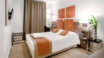 Quarto de hotel com uma cama, escrivaninha, cadeira, mesa de cabeceira, luminária e um vaso com flores.