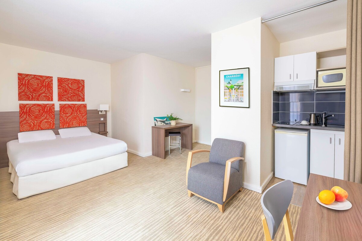 Habitación de hotel con cama, escritorio, silla, una pequeña mesa con fruta y kitchenette.