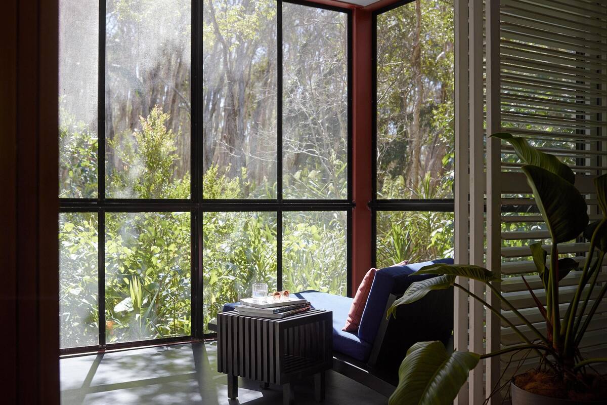 Ein Zimmer mit großem Fenster und Blick auf einen üppigen Garten. Auf einem kleinen Tisch stehen Bücher und eine Sonnenbrille.