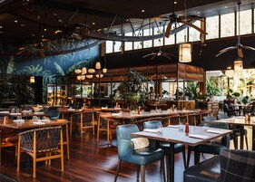 Un restaurante con mesas y sillas de madera, ventanas amplias y lámparas colgantes.