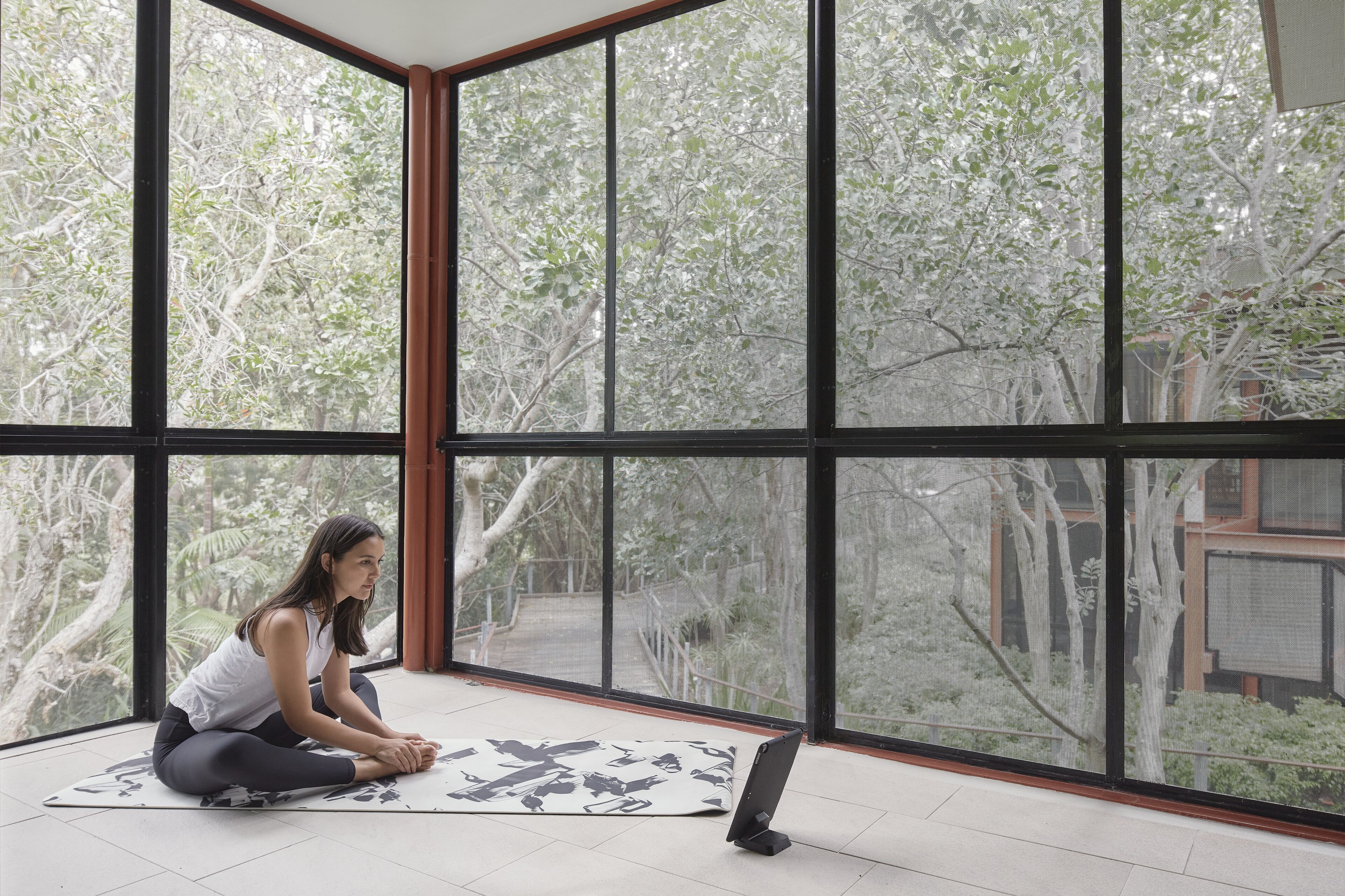 A person sitting on a mat in a room with large windows overlooking a garden.