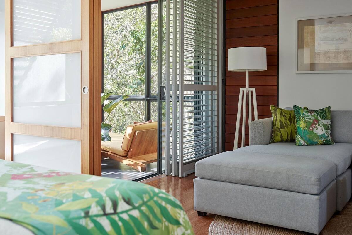 Ein Schlafzimmer mit einem Bett, einem grauen Sofa, einer Stehlampe und einem Fenster mit Jalousien.