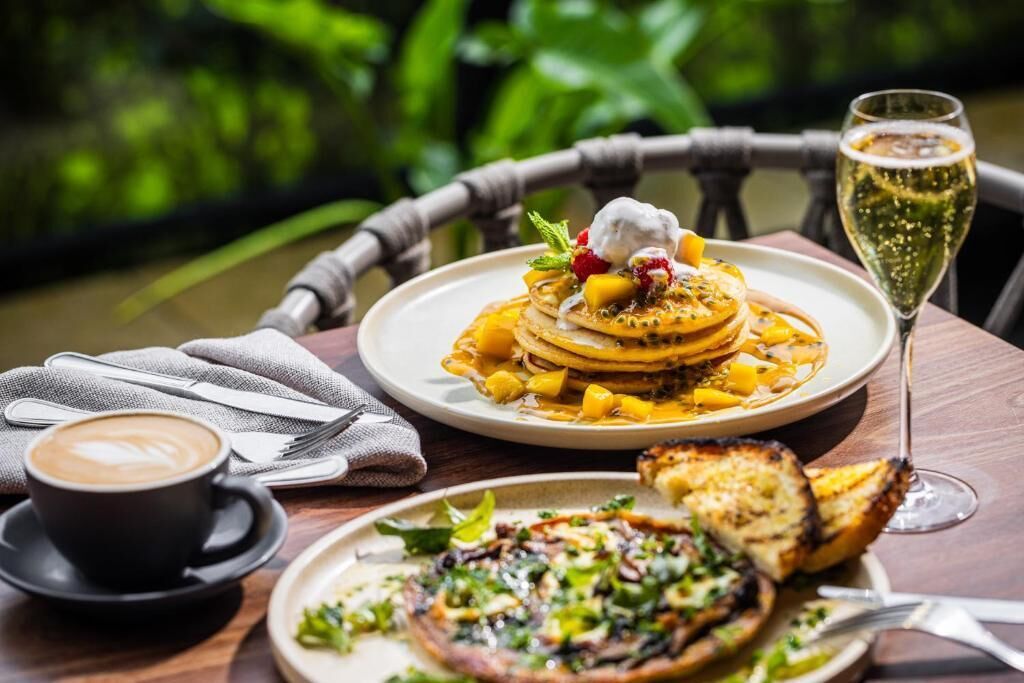 A dining table with a plate of pancakes topped with fruit and a dollop of cream, a glass of champagne, a cup of coffee, and a plate of grilled vegetables.