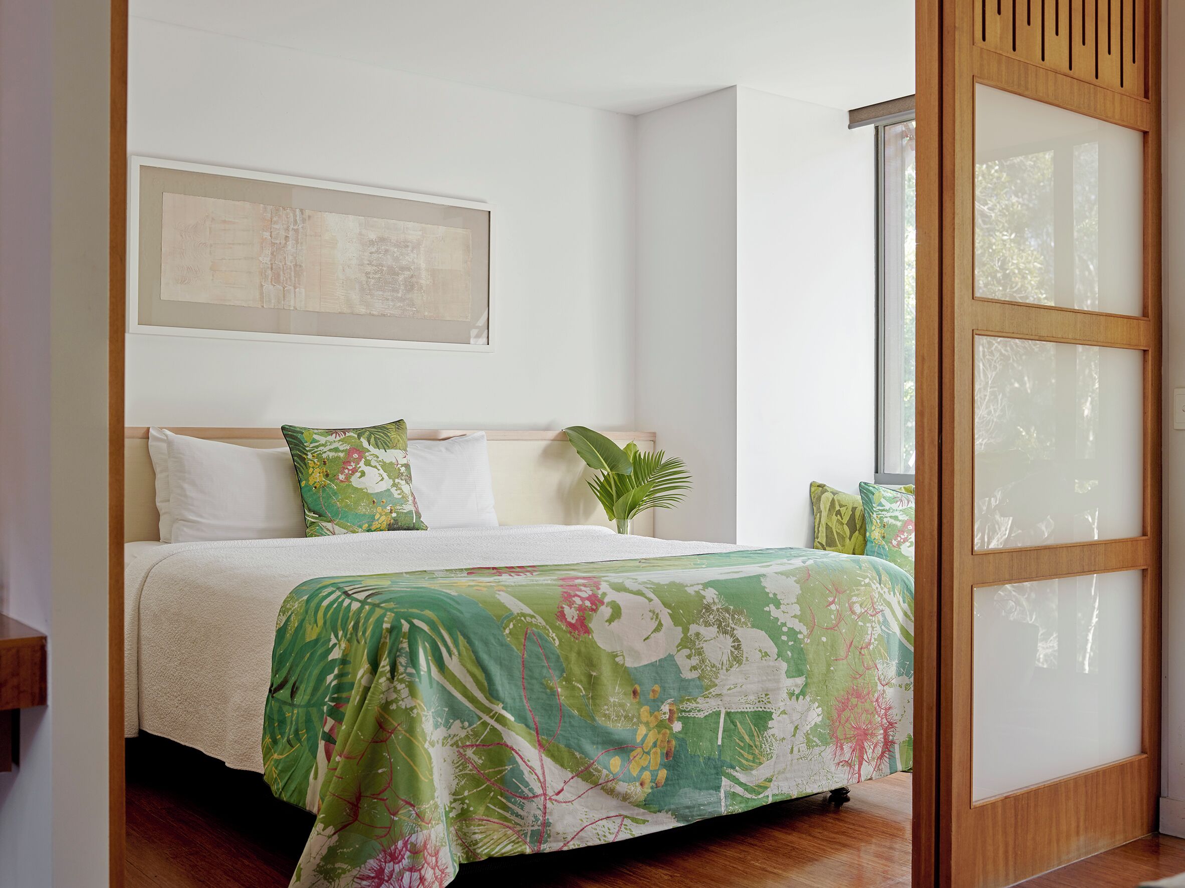 A neatly made bed with a floral bedspread, a large window with a wooden frame, and a wall-mounted artwork.