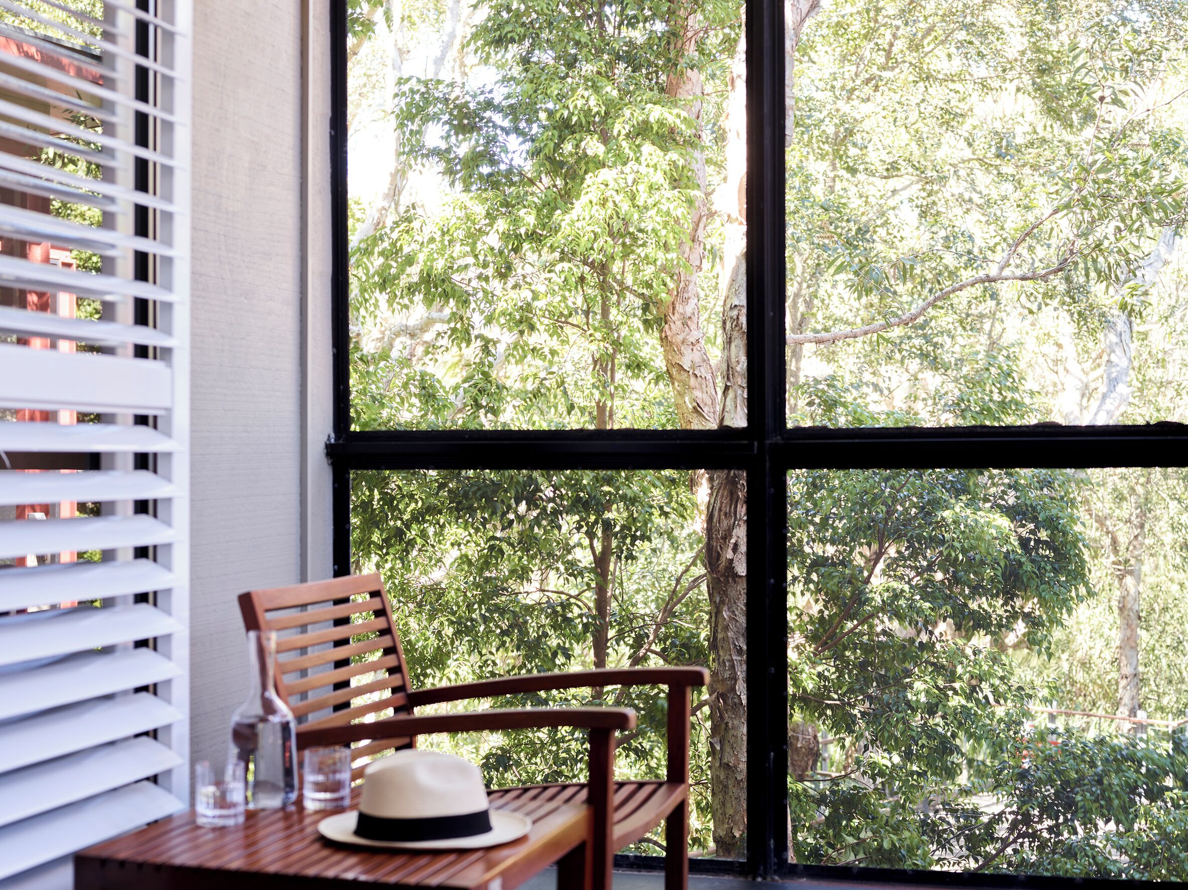 A wooden chair with a hat and glass on a table by a window with blinds.