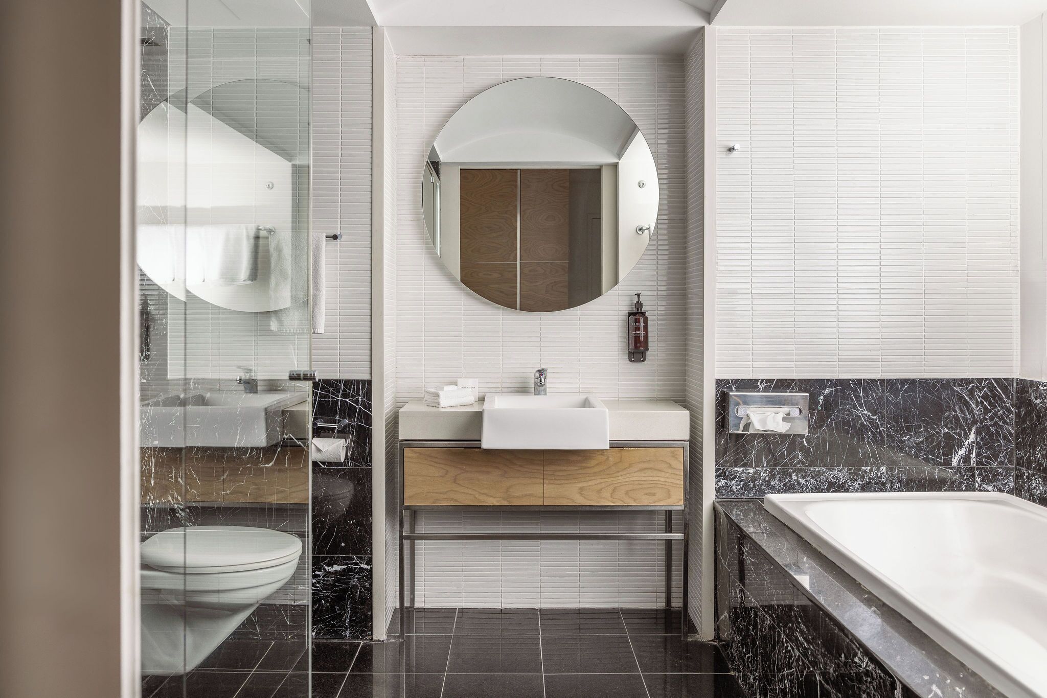 A modern bathroom with a glass shower enclosure, a round mirror, a wooden vanity, a white sink, and a marble-tiled bathtub.