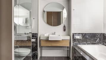Un baño moderno con cabina de ducha de vidrio, un espejo redondo, tocador de madera, lavamanos blanco y bañera con azulejos de mármol.
