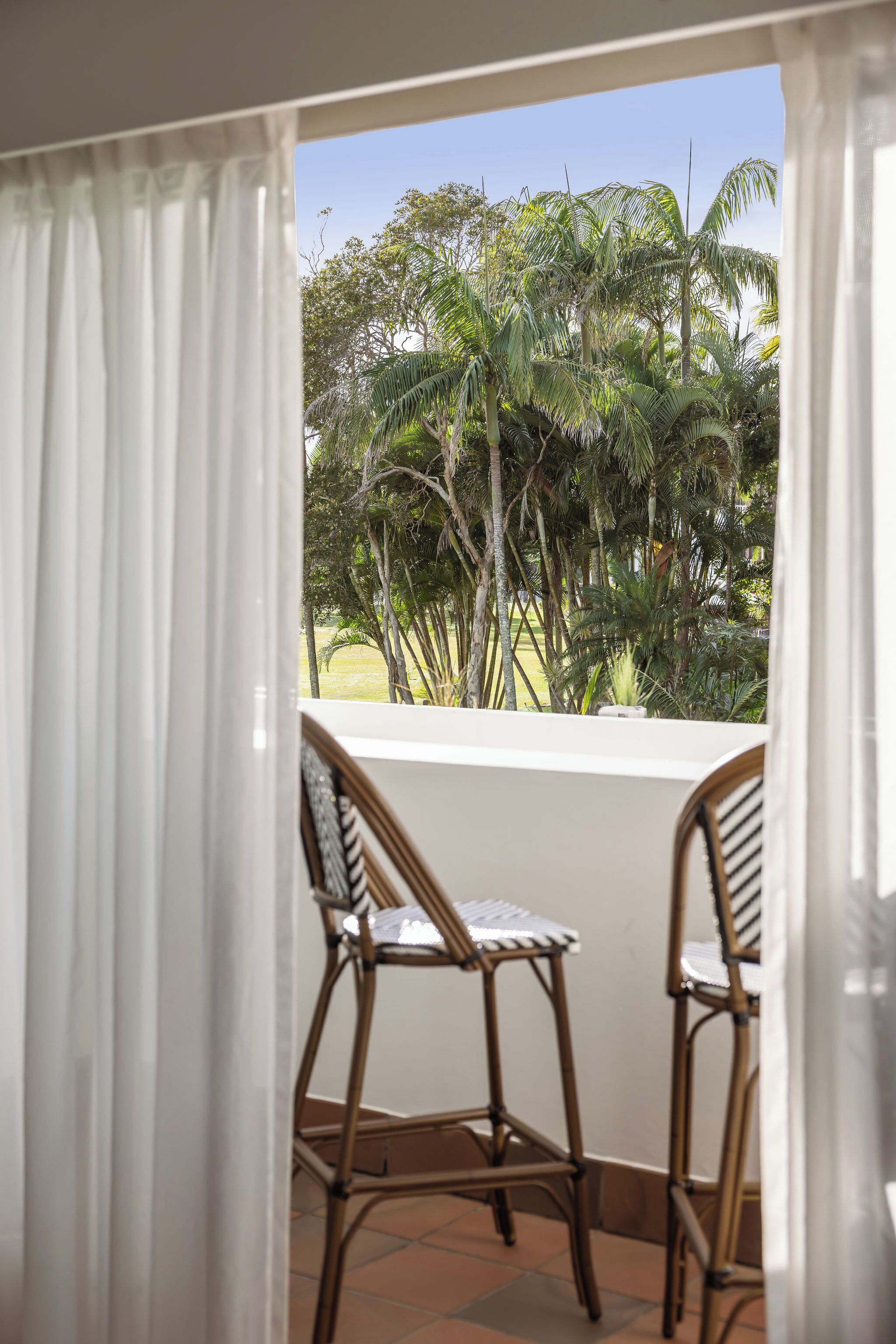 A balcony with two wicker chairs, white curtains, and a view of palm trees.
