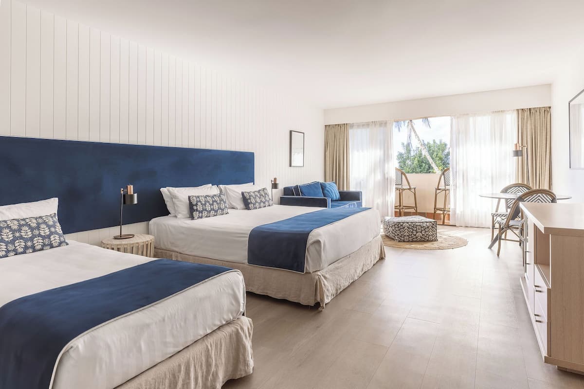 A hotel room with two beds, a desk, and a balcony with chairs.