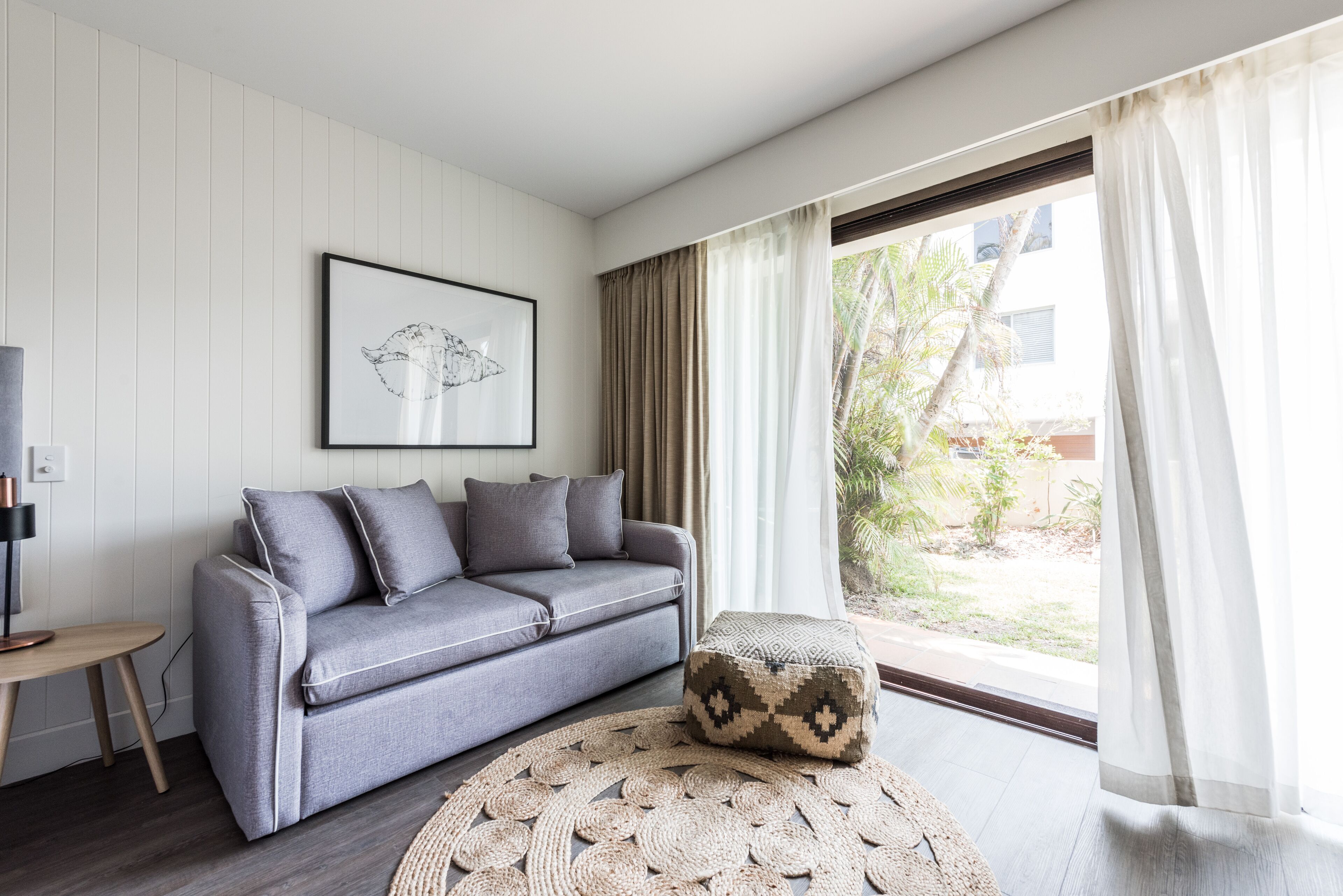 Un salon moderne avec un canapé gris, un pouf à motifs, un tapis rond en fibres tressées, une œuvre d’art encadrée accrochée au mur et une grande fenêtre avec des rideaux.
