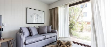 A modern living room with a grey sofa, a patterned ottoman, a round woven rug, a framed artwork on the wall, and a large window with curtains.