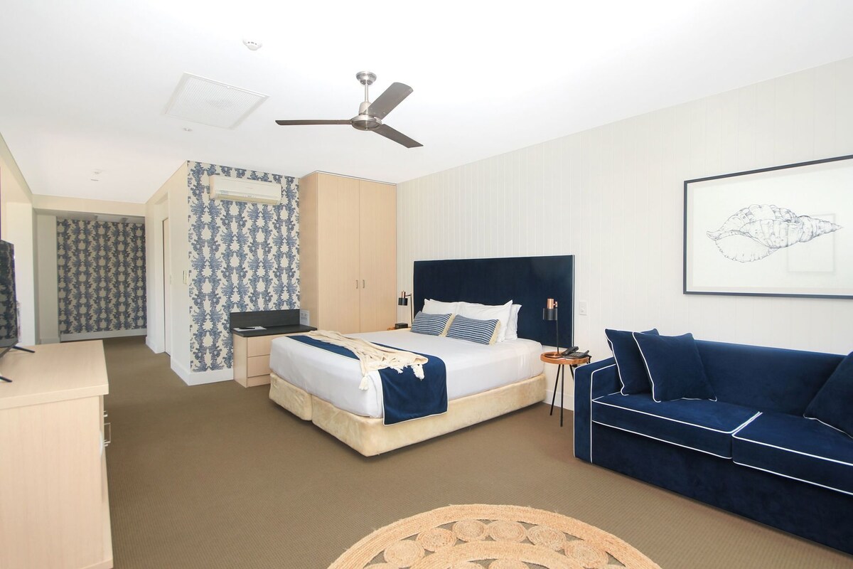A hotel room with a bed, a desk, a blue sofa, a ceiling fan, and a framed artwork on the wall.