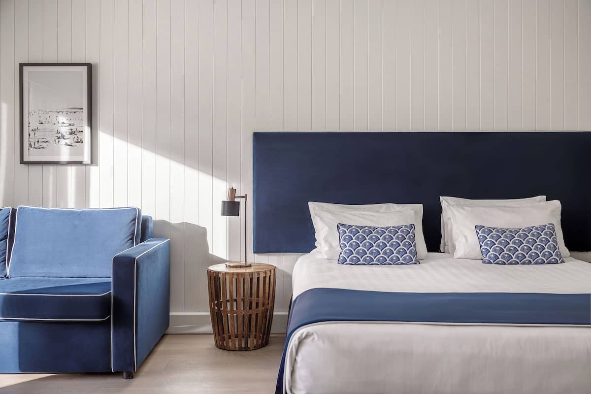A bedroom with a bed, a blue sofa, a small wooden side table, and a framed picture on the wall.