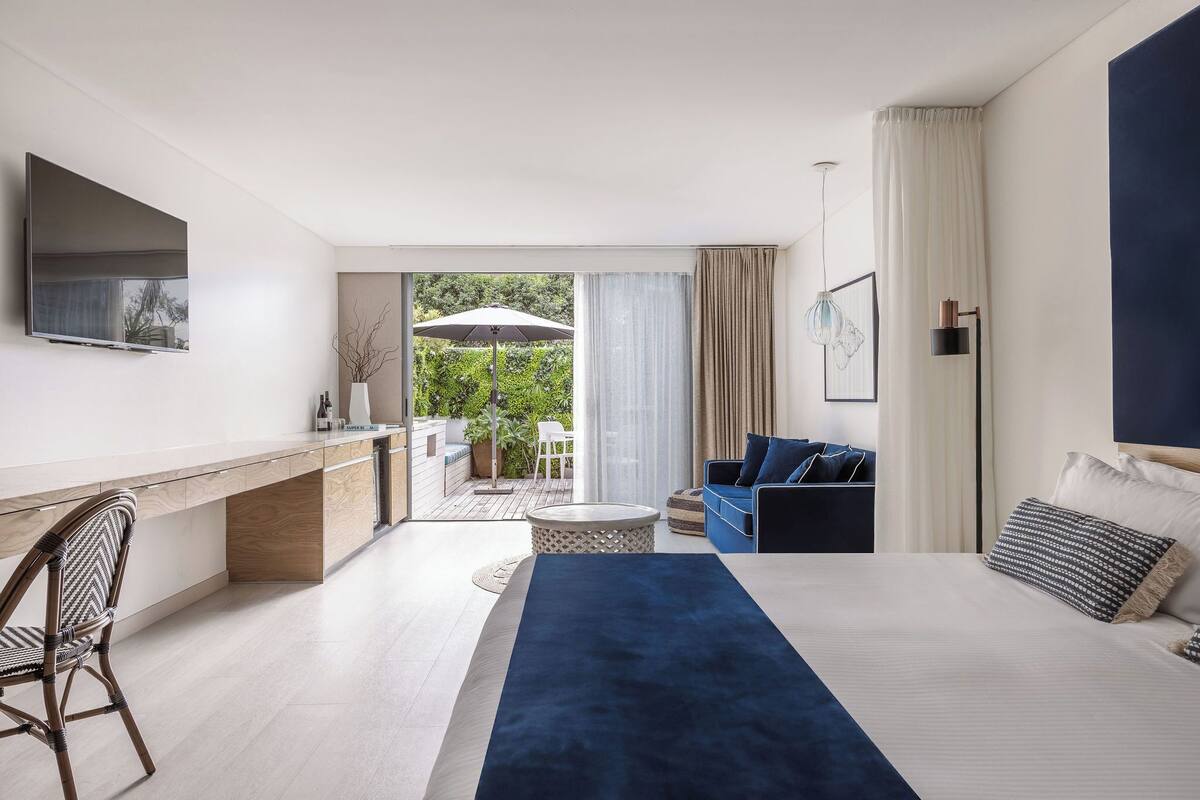 A modern hotel room with a large bed, a desk, a sofa, and a view of the outdoors.