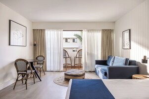 Habitación de hotel con cama, sofá, mesa de comedor con sillas y balcón con cortinas.