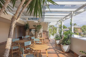 Un espacio al aire libre cubierto con mesas y sillas de madera, plantas en macetas y vistas del barrio circundante.