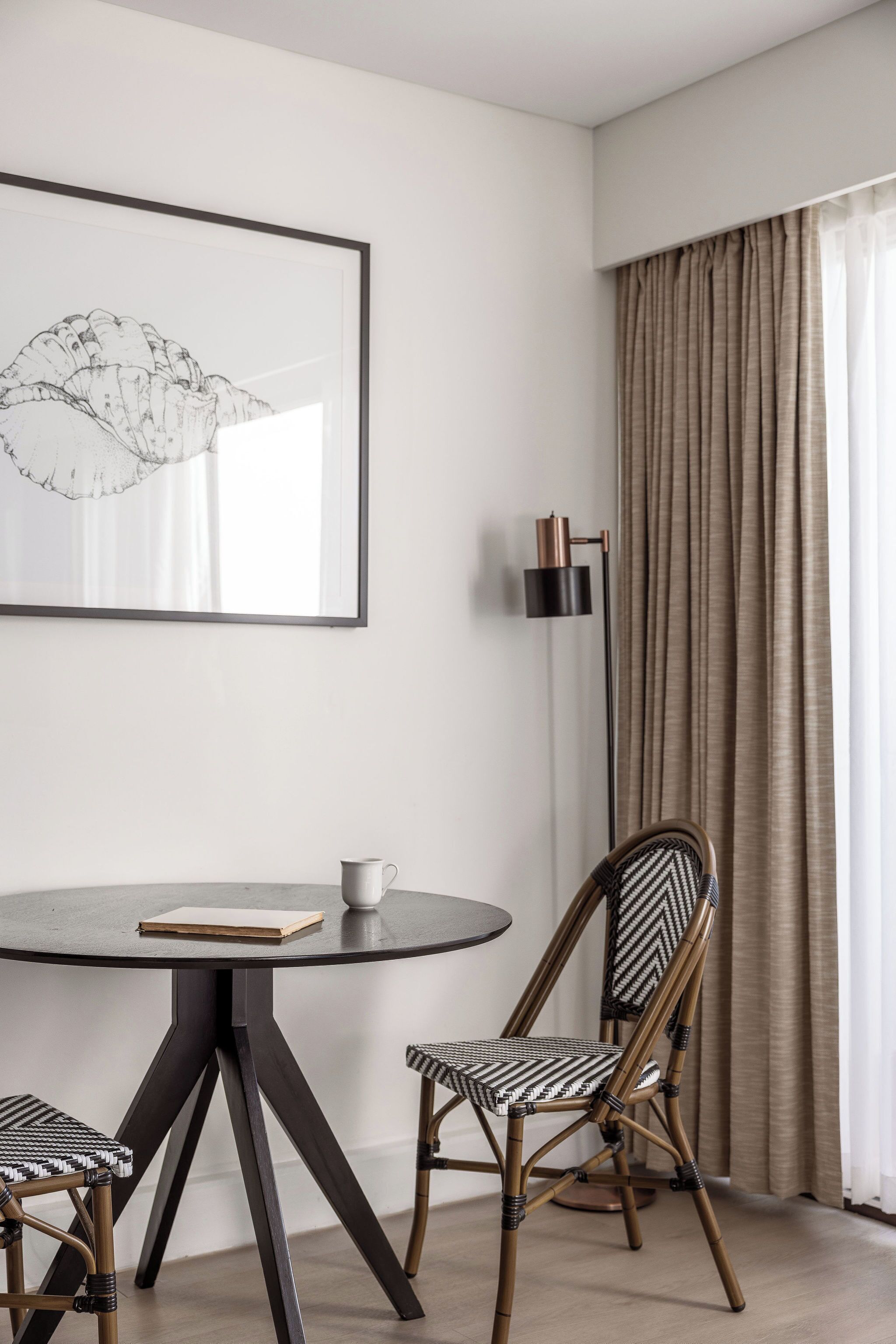 Une pièce moderne avec une table ronde, deux chaises, une œuvre d’art encadrée, un lampadaire et une fenêtre avec des rideaux.