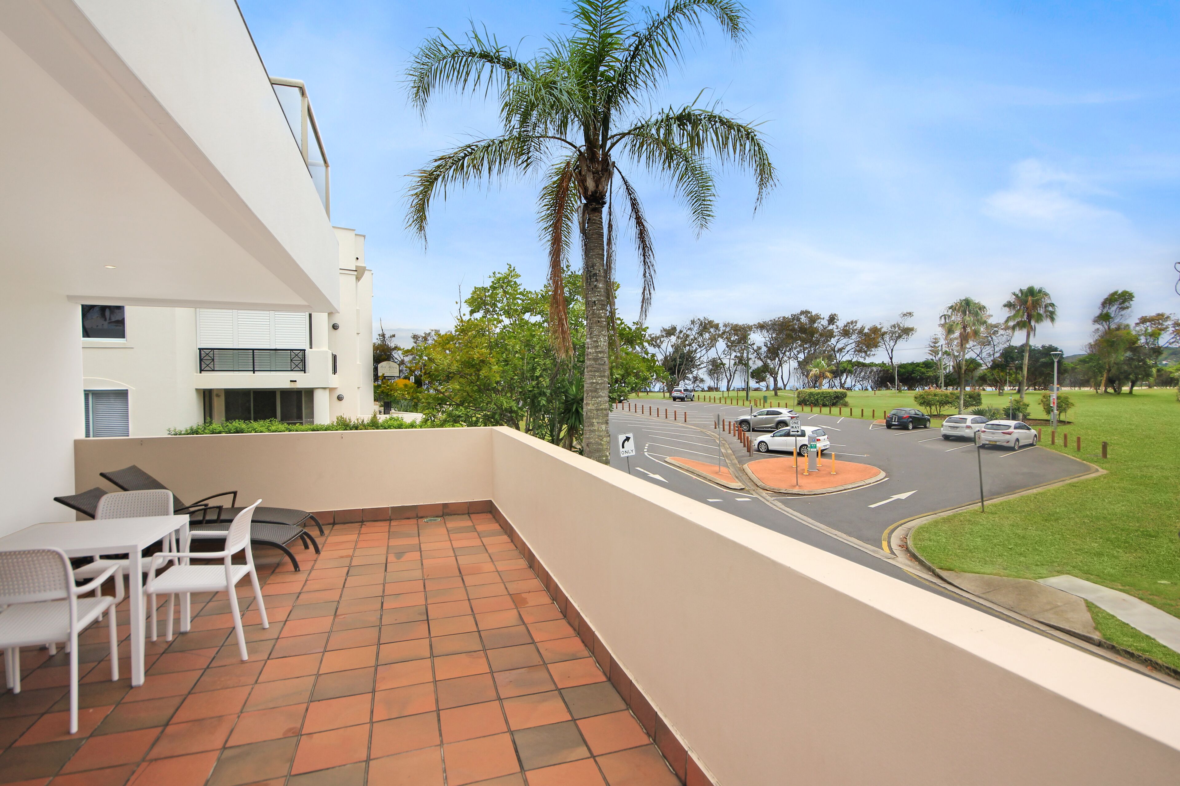 A balcony with a tiled floor, white furniture, and a view of a street with cars and palm trees.