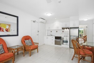 Una cocina moderna con armarios blancos, horno empotrado y refrigerador. Hay sillas de mimbre con cojines naranja y una mesa de vidrio.
