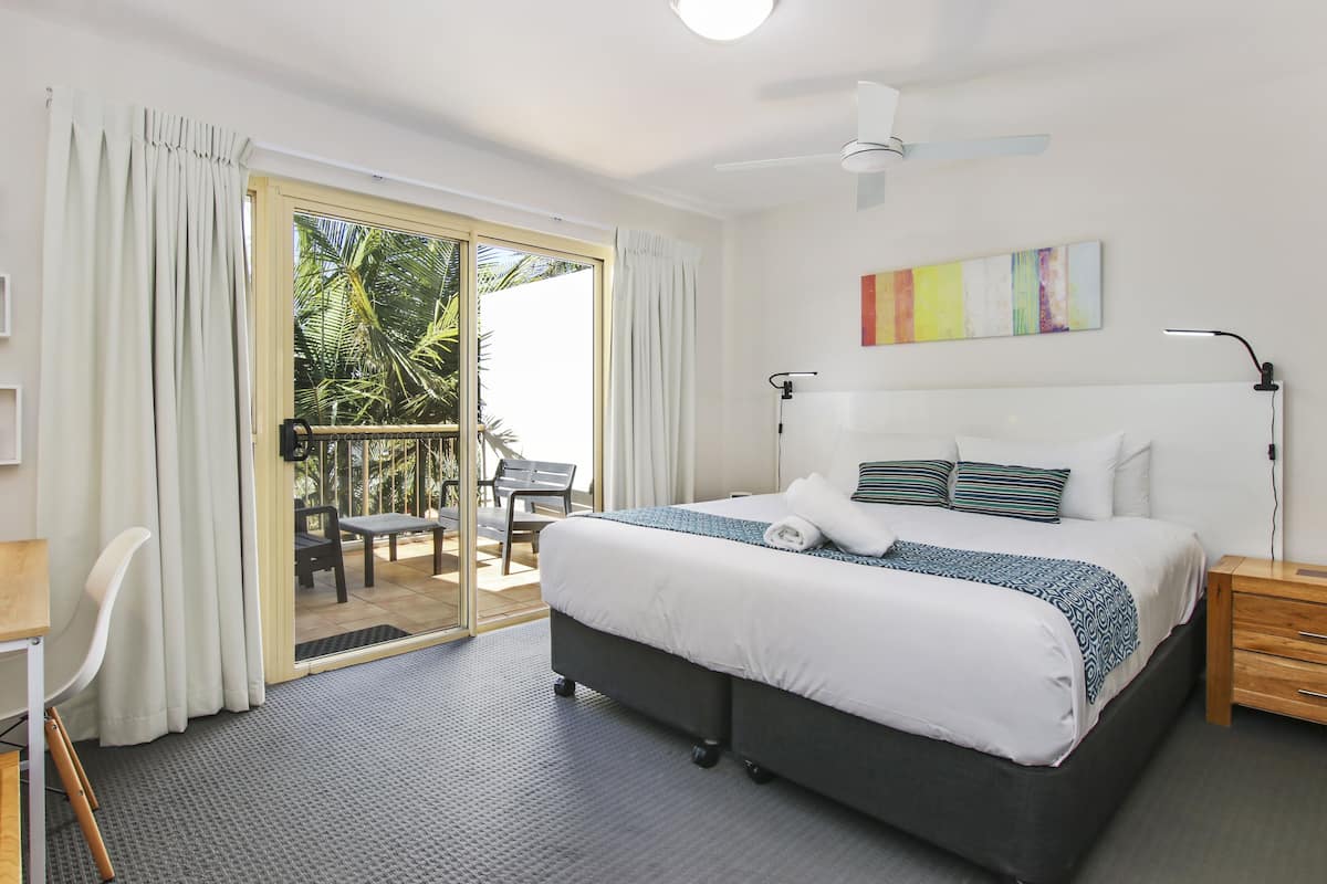 Ein Hotelzimmer mit Bett, Schreibtisch, Stuhl, Nachttisch, Lampe, Deckenventilator und einer Glastür, die zu einem Außenbereich mit Tisch und Stühlen führt.