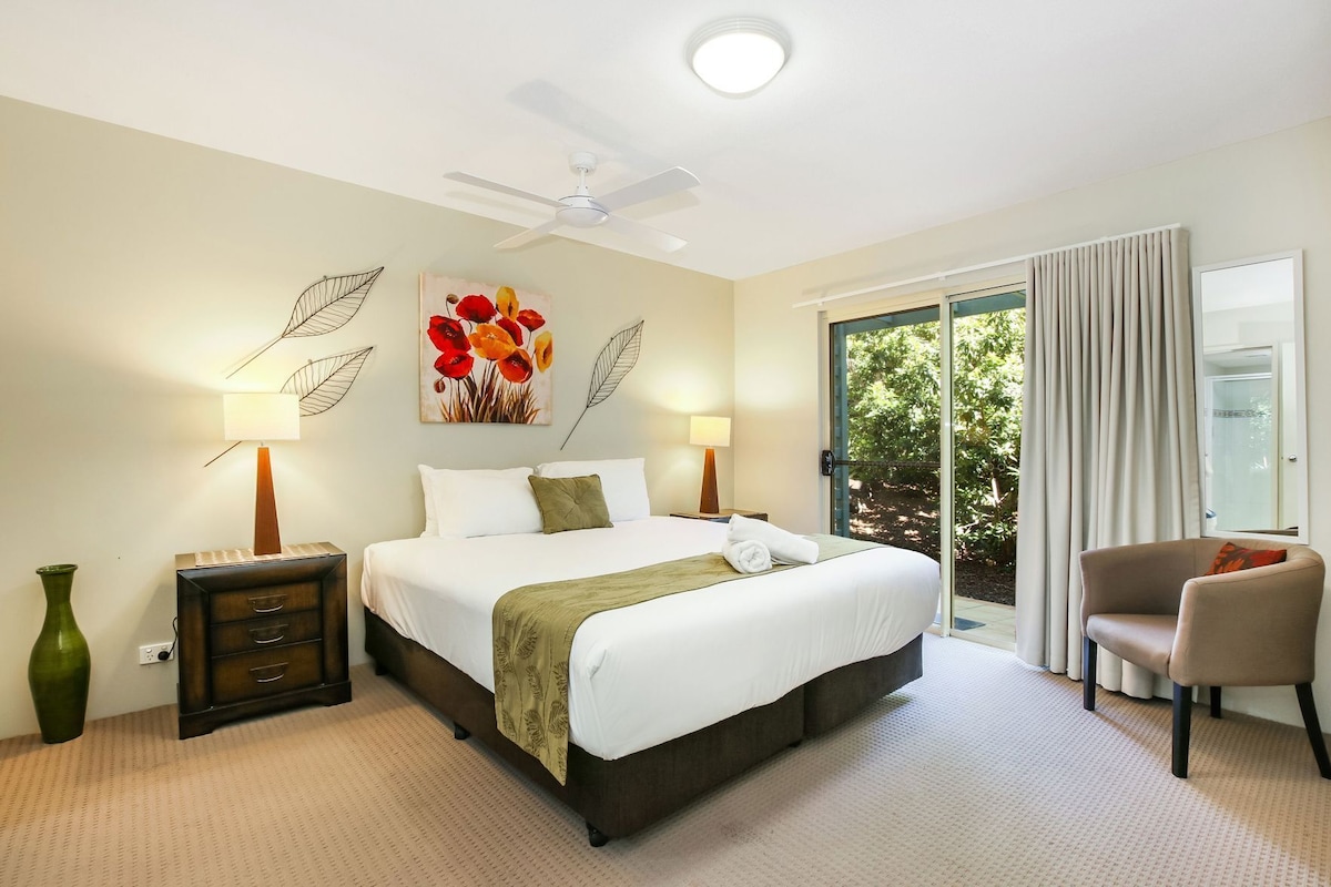 A hotel room with a bed, bedside lamps, a green vase, a chair, and a view of greenery outside.