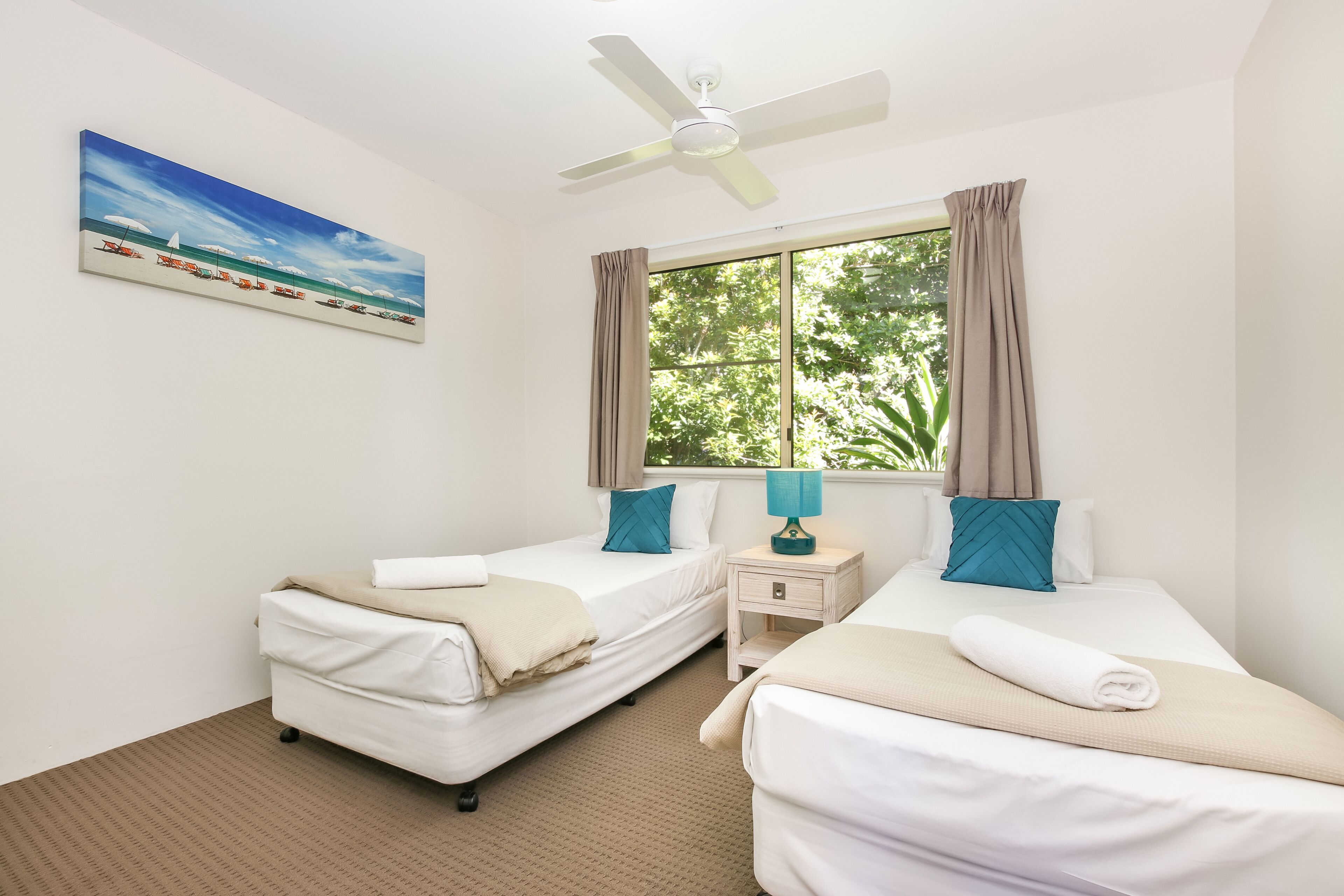A hotel room with two beds, a ceiling fan, a window with curtains, a nightstand with a lamp, and a beach-themed painting on the wall.