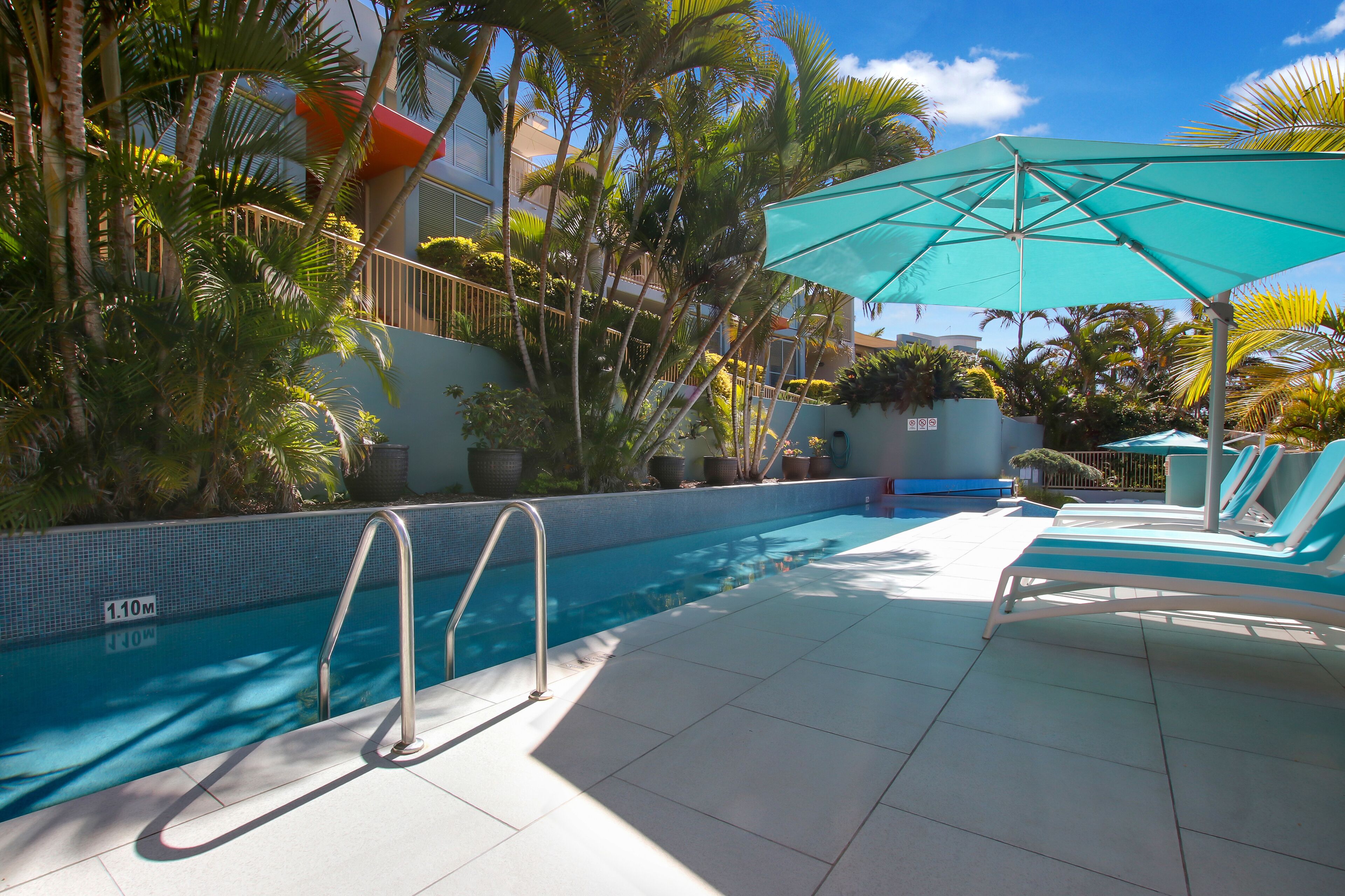 Une piscine dont la profondeur est indiquée à 1,10 mètre, entourée de palmiers et agrémentée d’un espace de détente avec un parasol turquoise.