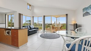 A modern kitchen with a glass dining table and white chairs, a black sofa, and a view of the outdoors through large windows. - Lennox Beach Resort (Lennox Head)